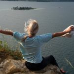 Woman enjoys a breathtaking lake view from a cliff at sunset, exemplifying freedom and adventure.